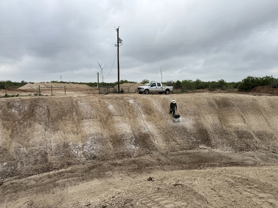 A dusty, barren landscape with a pickup truck and construction equipment, including wind turbines in the background, and a lone worker walking across the terrain.