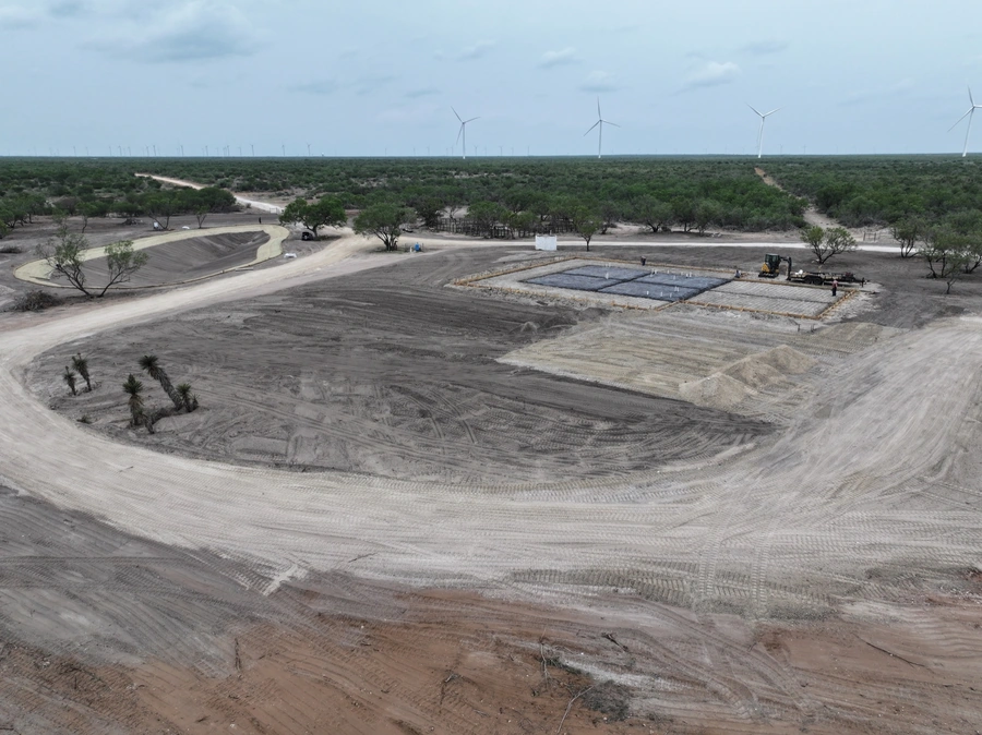 Aerial view of a large construction site in a rural, open area with wind turbines visible in the background. The site shows significant earthmoving and grading work underway, with several areas of exposed soil and gravel.