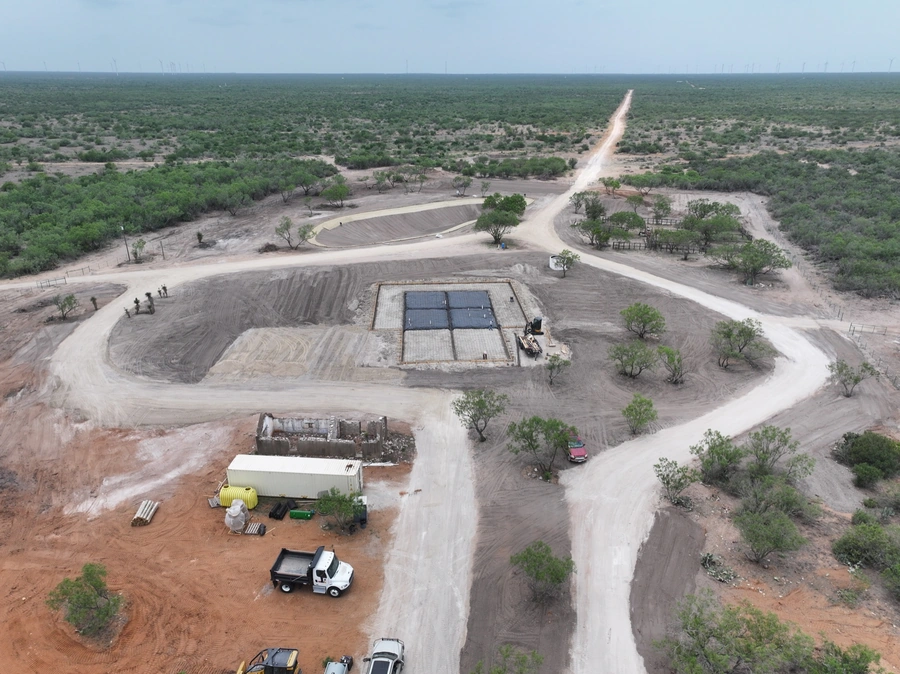 Aerial view of a construction site in a rural, remote area. The site includes a large equipment shed, trucks, and various construction materials scattered around the site. The surrounding landscape is dominated by scrubland and dirt roads, with few trees visible.