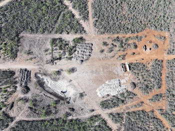 Aerial view of a construction site in a forested area, with heavy machinery, excavated earth, and piles of materials visible.