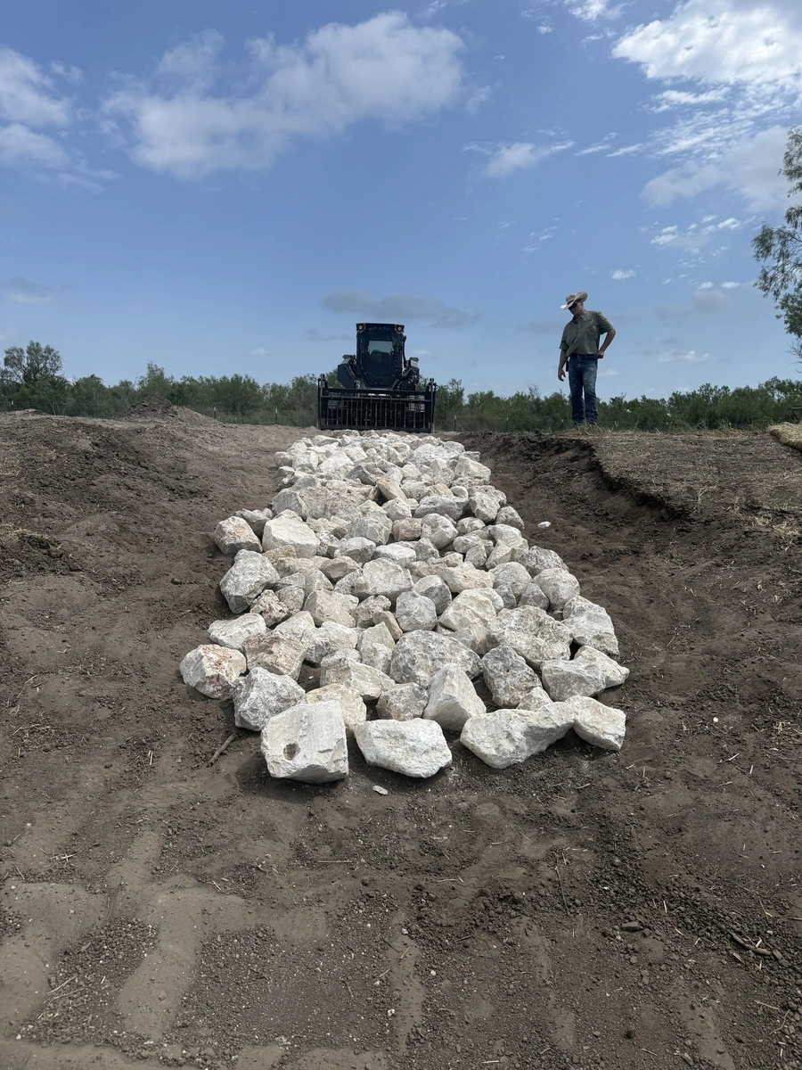 A construction site with a large pile of white rocks and a backhoe in the background. A worker is standing next to the rock pile.