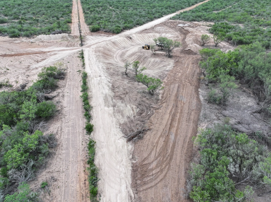 Aerial view of a large construction site in a rural, wooded area. A heavy machinery vehicle is visible on the cleared, dusty path cutting through the landscape.