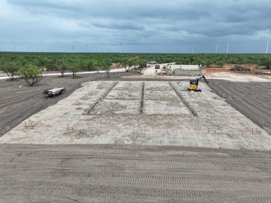 Aerial view of a construction site with heavy machinery, dirt mounds, and wind turbines in the background on a cloudy day.