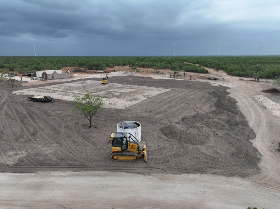 Aerial view of a construction site in a rural, wind-powered environment. Large construction equipment, including a bulldozer, is visible, indicating land clearing and site preparation work underway. The site is surrounded by green vegetation and wind turbines, suggesting a remote or industrial setting.