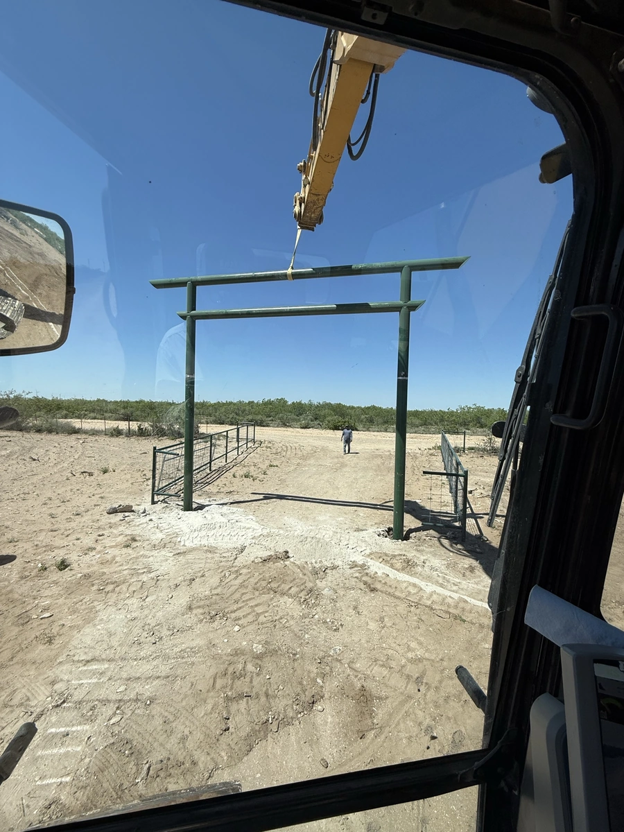 A heavy construction crane lifting a metal frame for a gate or entrance structure in a remote, arid landscape with a person walking in the distance.