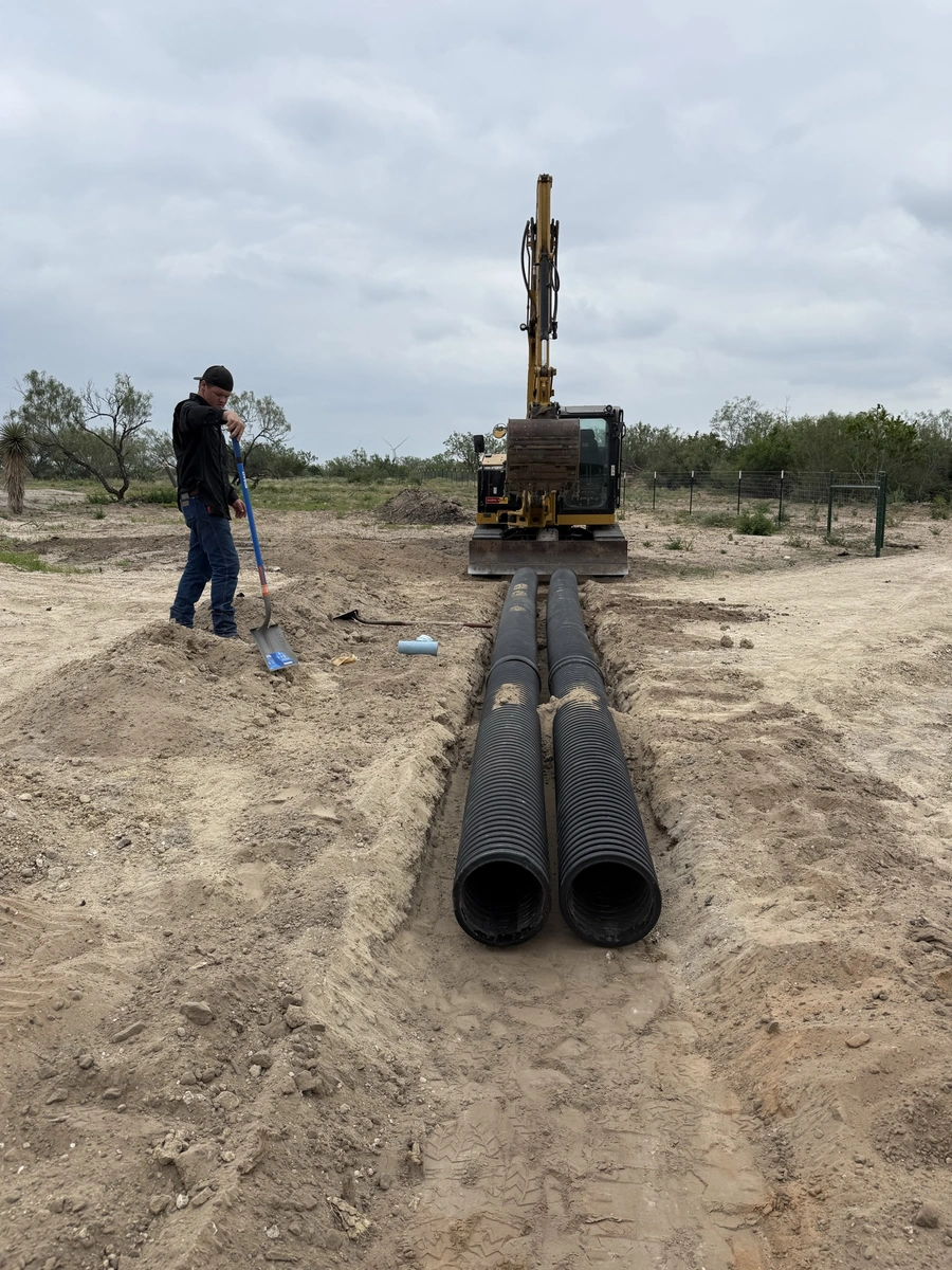 A construction worker operating a large excavator machine as part of a land improvement project. The excavator is digging a trench in a dusty, rural landscape, with pipes laid out ready for installation.
