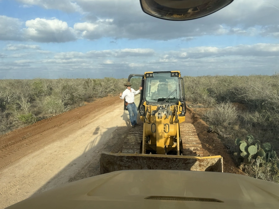 A construction worker operating a yellow backhoe tractor on a dirt road surrounded by sparse vegetation and cloudy sky in a rural setting.
