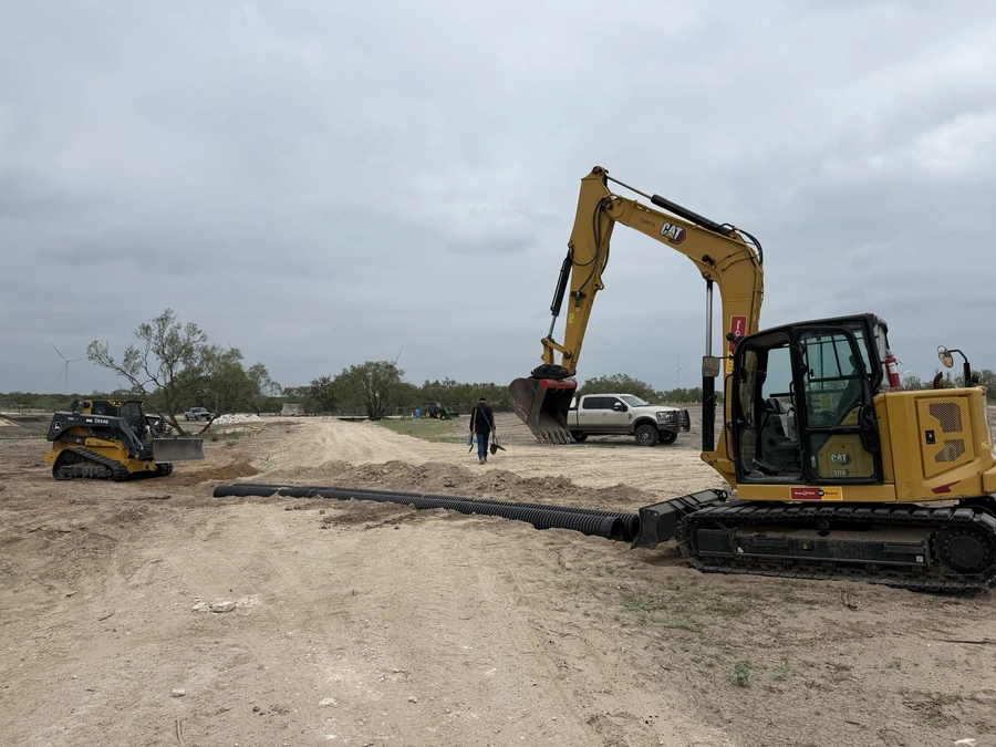 A construction site with heavy machinery and equipment, including an excavator, bulldozer, and various other vehicles. The site appears to be undergoing significant earthwork or site preparation, with piles of dirt and a cleared, open area visible.