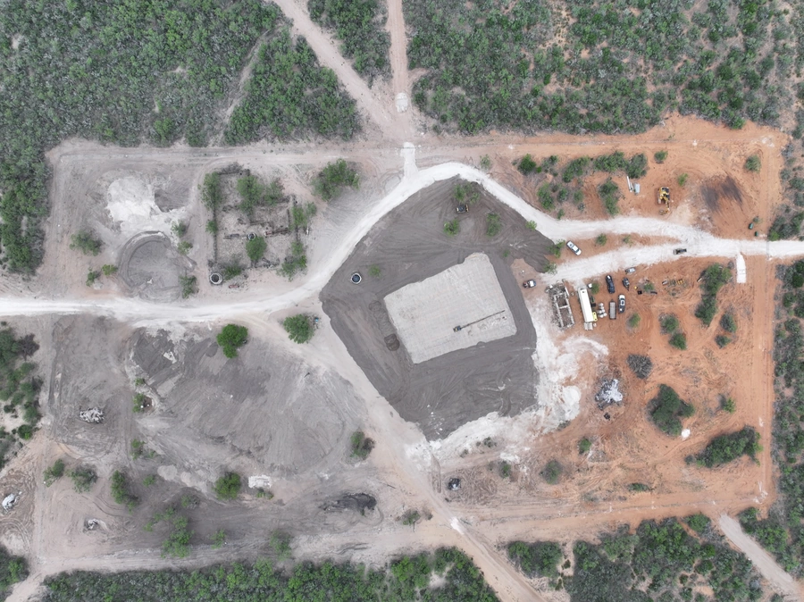 Aerial view of a construction site with heavy machinery, piles of soil, and a large building under construction surrounded by wooded areas.