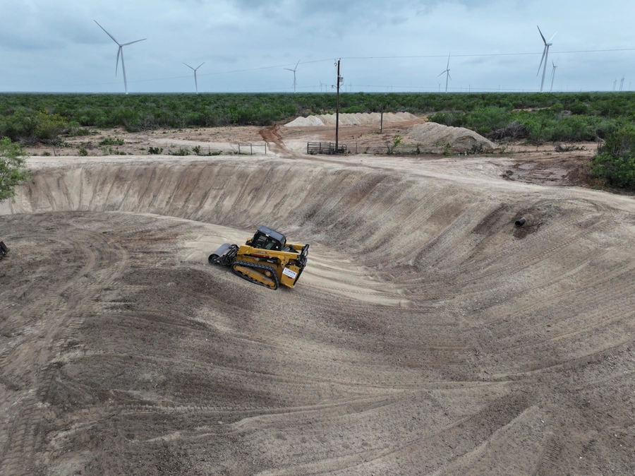 A large yellow construction vehicle on a dirt construction site with wind turbines in the background, indicating a renewable energy project.