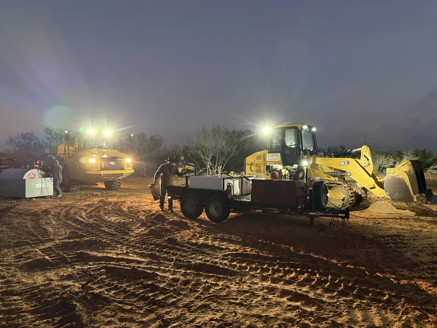 Construction equipment and workers are shown working on a nighttime project in a rural area. Several large construction vehicles, including a backhoe and dump truck, are illuminated by bright lights and operate on a dirt and gravel surface. The scene depicts the active work of A.M. Pitts Construction, a company that specializes in land improvement, ranch work, site work, excavation, and utility services throughout Central Texas.