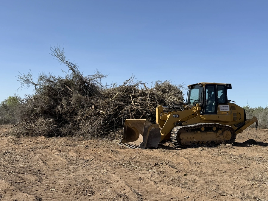 A large bulldozer/crawler tractor moving through a field, pushing a large pile of branches and debris.