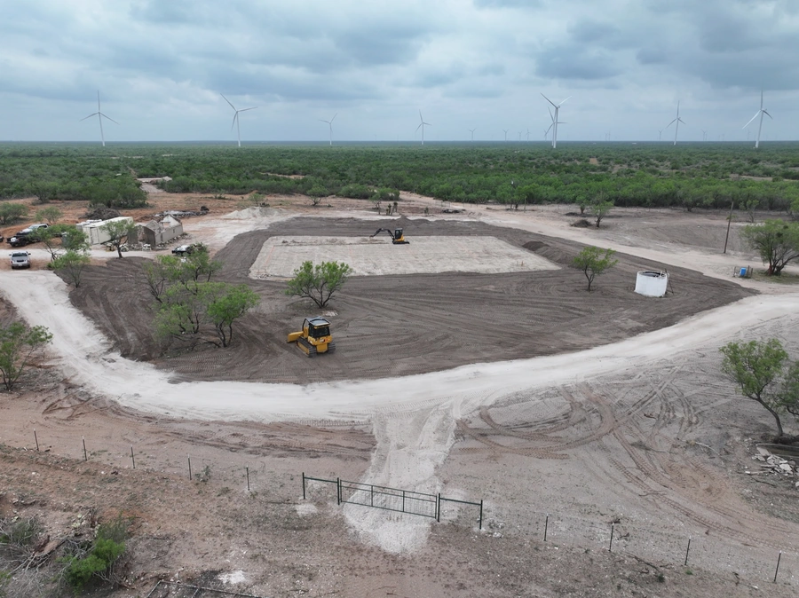 A large construction site with heavy machinery, wind turbines in the background, and an expansive natural landscape.