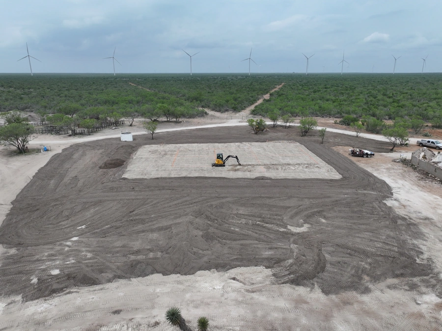 Aerial view of a large construction site with heavy equipment and wind turbines visible in the background. The site appears to be undergoing significant land preparation and earthmoving work.