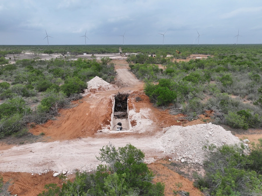 An aerial view of a construction site in a rural, forested area. The image shows a winding dirt road through the brush, with several large wind turbines visible in the background. The road appears to be under construction, with piles of dirt and debris visible.