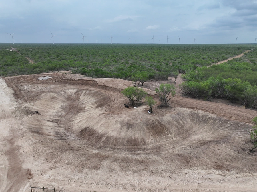 Aerial view of a large construction site in a grassy, rural area with wind turbines in the distance. The site appears to be undergoing major land clearing and earthmoving work, with deep trenches and piles of excavated soil visible.