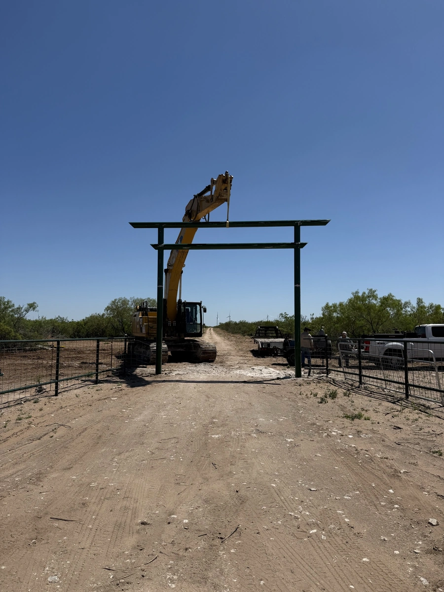 A construction site with a large excavator lifting a metal framework, surrounded by fencing and equipment. The image suggests ongoing land development or site preparation work.