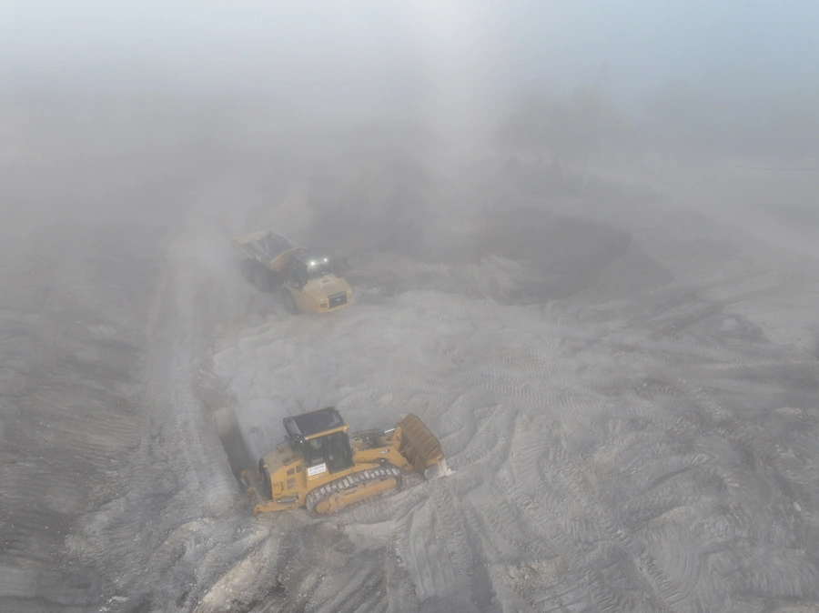 Aerial view of two bulldozers operating in a foggy, dusty construction site surrounded by rugged terrain and natural vegetation.