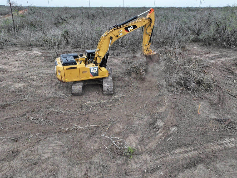 A yellow Caterpillar excavator machine digging into the dirt and clearing brush in a remote, rural landscape with sparse vegetation and a dusty, uneven terrain.