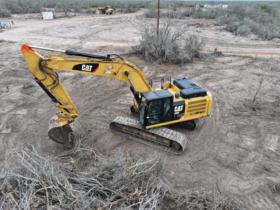 A large yellow Caterpillar excavator moving soil and debris at a construction site in a rural, scrubby landscape. The excavator's boom is extended and the bucket is lowered to the ground, indicating it is actively engaged in moving material. A road grader or other heavy equipment can be seen in the background, suggesting ongoing land clearing or site preparation work.
