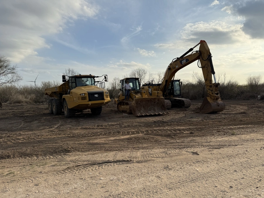 A construction site with several heavy machinery vehicles, including a dump truck and excavators, working on a dirt clearing project under a partly cloudy sky.