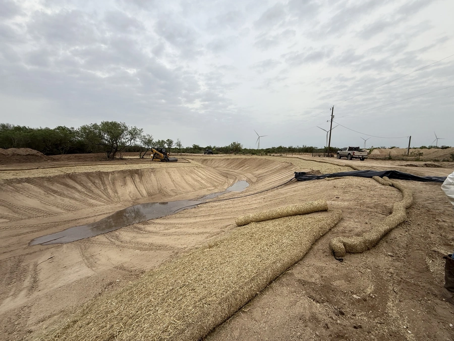 A construction site with heavy equipment and materials laid out for a large excavation and land improvement project. The image shows a vast, sandy field with tire tracks, piles of gravel, and wind turbines in the background, indicating a rural, open environment.