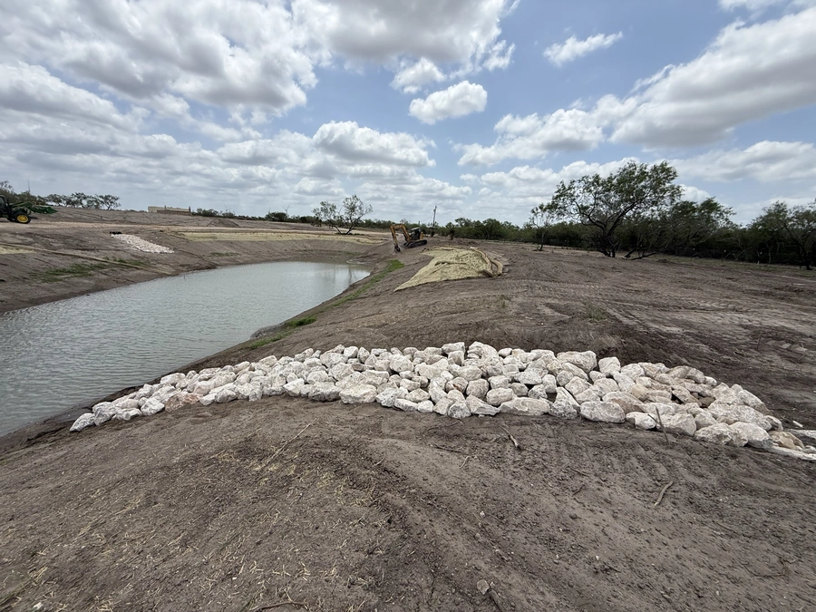 Landscape view of an excavation site with a pond, heavy machinery, and a pile of crushed rock. The site appears to be undergoing land improvement or construction work.