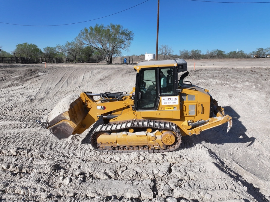 A yellow bulldozer tractor working on a construction site, clearing and grading the land. The tractor has tracks instead of wheels and a large front-mounted blade for moving soil and debris.