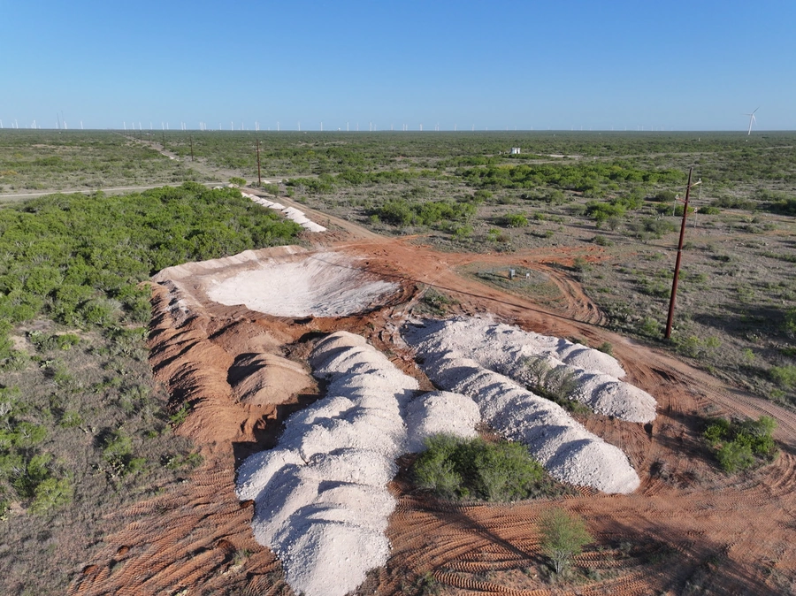An aerial view of a remote, rural landscape with a winding dirt road cutting through the scrubby vegetation. In the distance, rows of wind turbines can be seen on the horizon, indicating the area's use for renewable energy production.