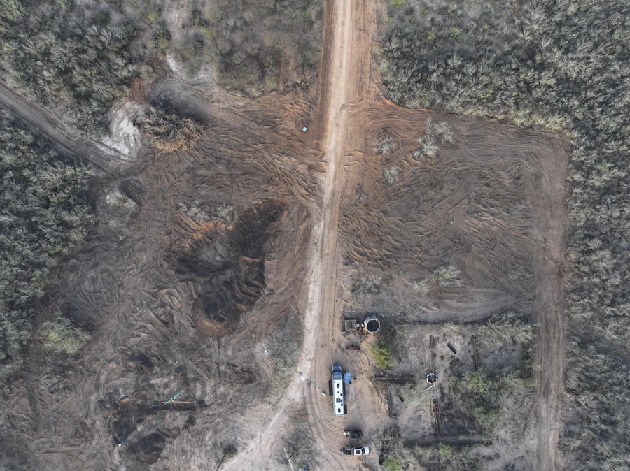 Aerial view of a large construction site in a wooded area, showing a long dirt road winding through the landscape with several trucks and equipment parked along the sides.