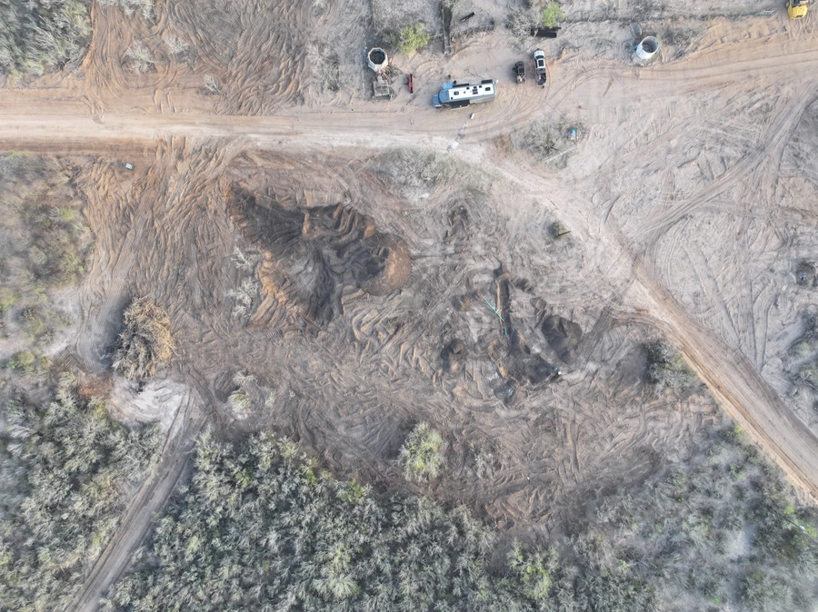 Aerial view of an active construction site, with heavy machinery, trucks, and equipment visible on a cleared and excavated plot of land surrounded by natural vegetation.