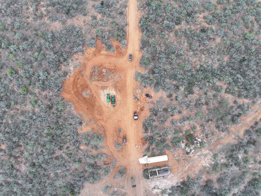 Aerial view of a remote construction site in a forested area, with several trucks and heavy machinery visible on a red-dirt road.
