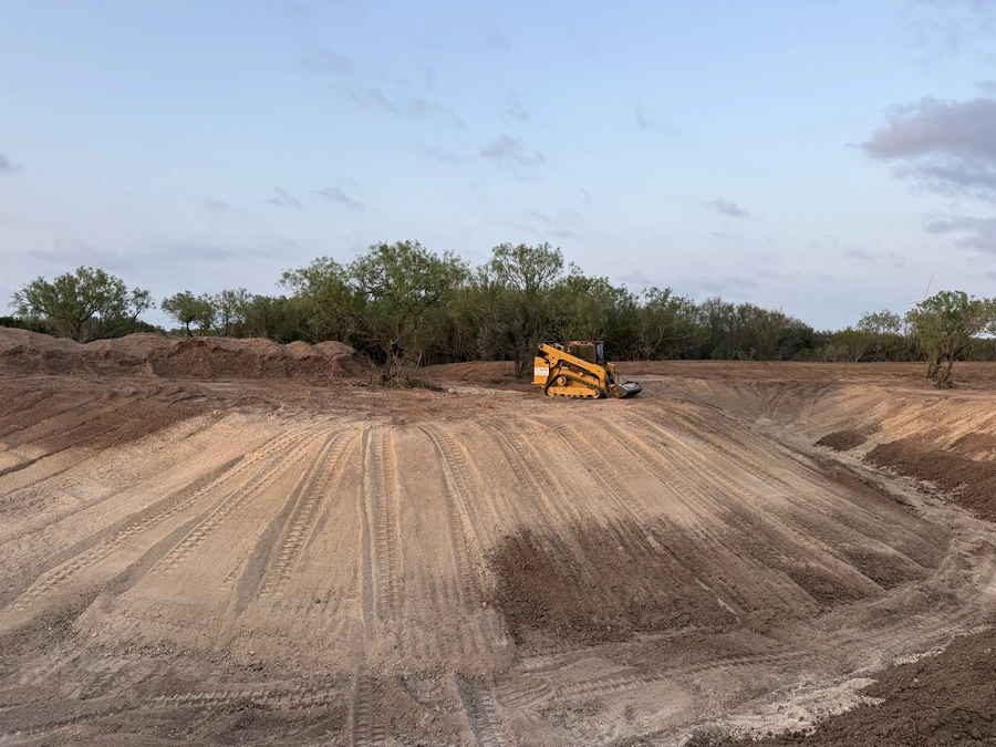 A large orange tracked skid steer excavator working in a barren, freshly graded field with piles of dirt and scattered trees in the background. The scene depicts land clearing and site preparation work for a construction project.