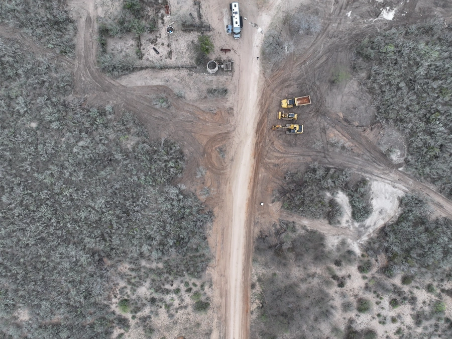 Aerial view of a construction site in a rural, wooded area. The site features a dirt road, heavy machinery like excavators and dump trucks, and various equipment and materials. The surrounding landscape is covered in dense vegetation, indicating a remote, natural setting.