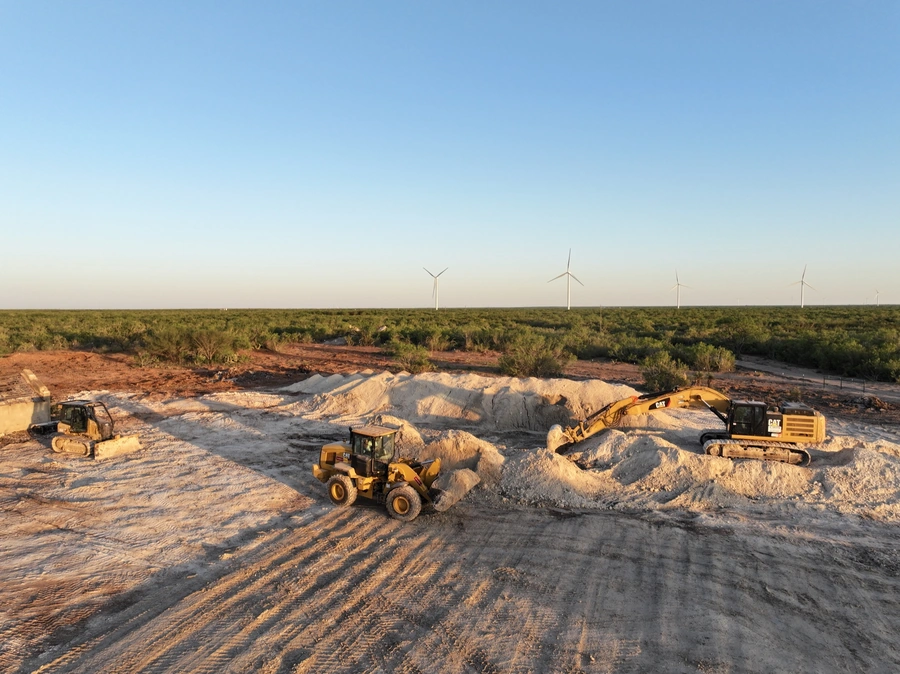 A construction site with heavy machinery, including bulldozers and excavators, actively shaping the terrain and clearing the way for the installation of a new wind farm. In the background, wind turbines can be seen, suggesting the project's renewable energy focus.