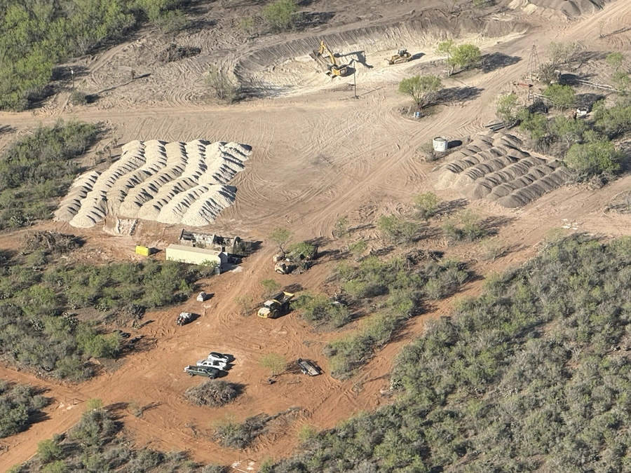 An aerial view of an active construction site in a rural, wooded area. The site shows heavy machinery, piles of dirt, and cleared land indicating ongoing land improvement and excavation work.