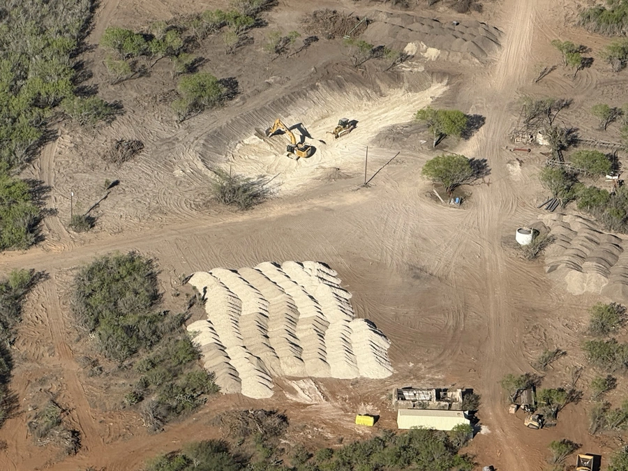 Aerial view of a construction site with heavy machinery and earthmoving equipment, such as an excavator, operating on a dirt and gravel surface with sparse vegetation. The site appears to be undergoing land clearing or site preparation work.
