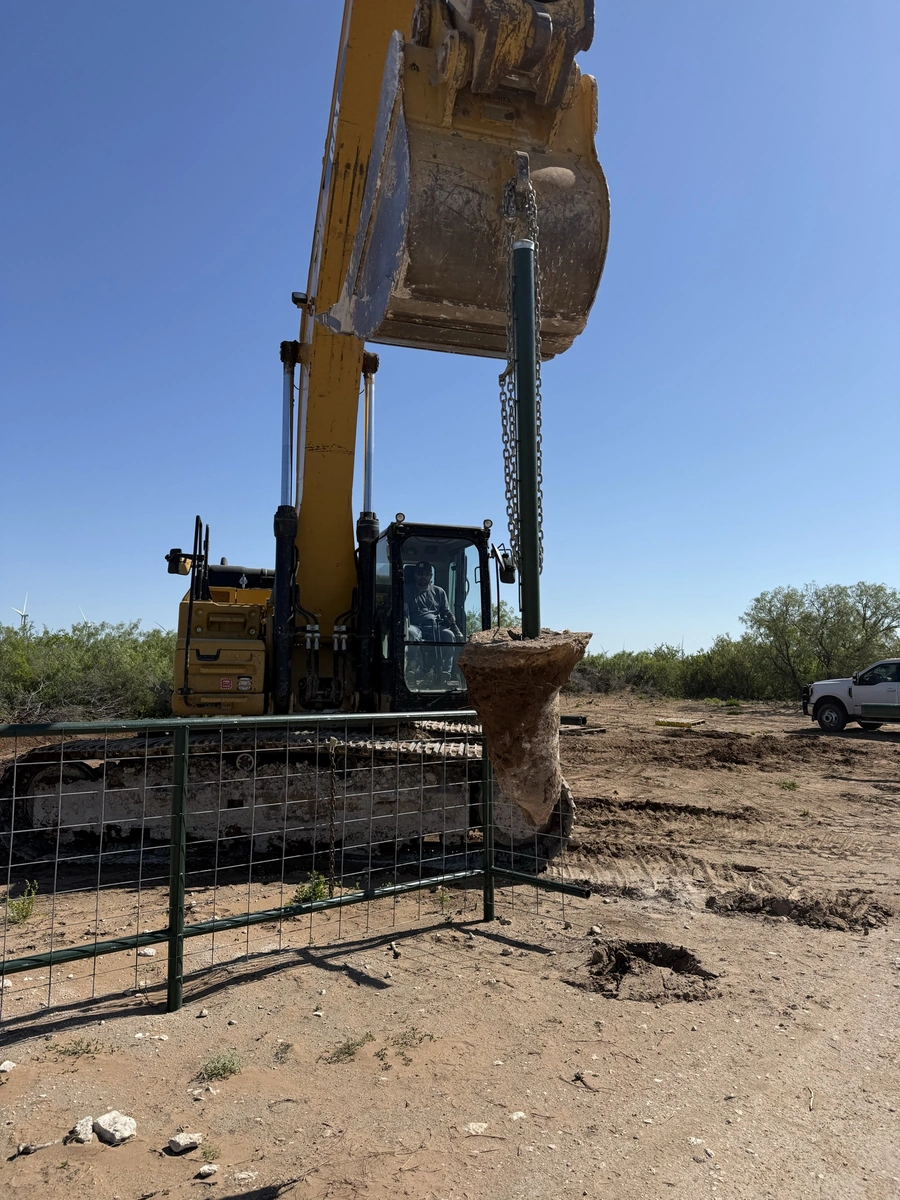 A large yellow excavator machine is shown digging into the ground on a remote construction site, surrounded by dirt, rocks, and a wire fence.