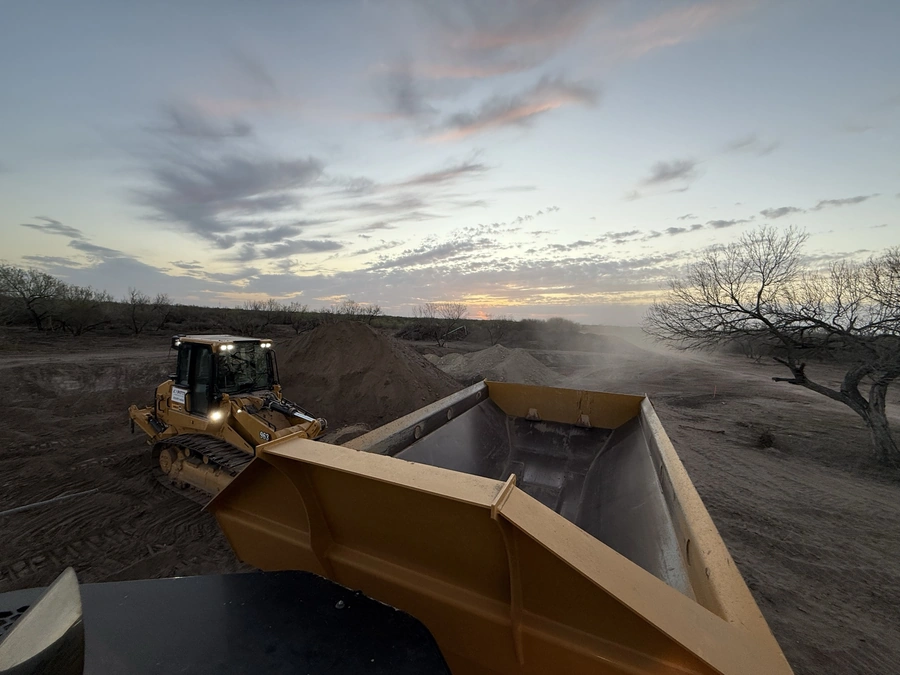 A bulldozer excavating soil at a construction site during sunset, with a hazy sky and silhouetted trees in the background.