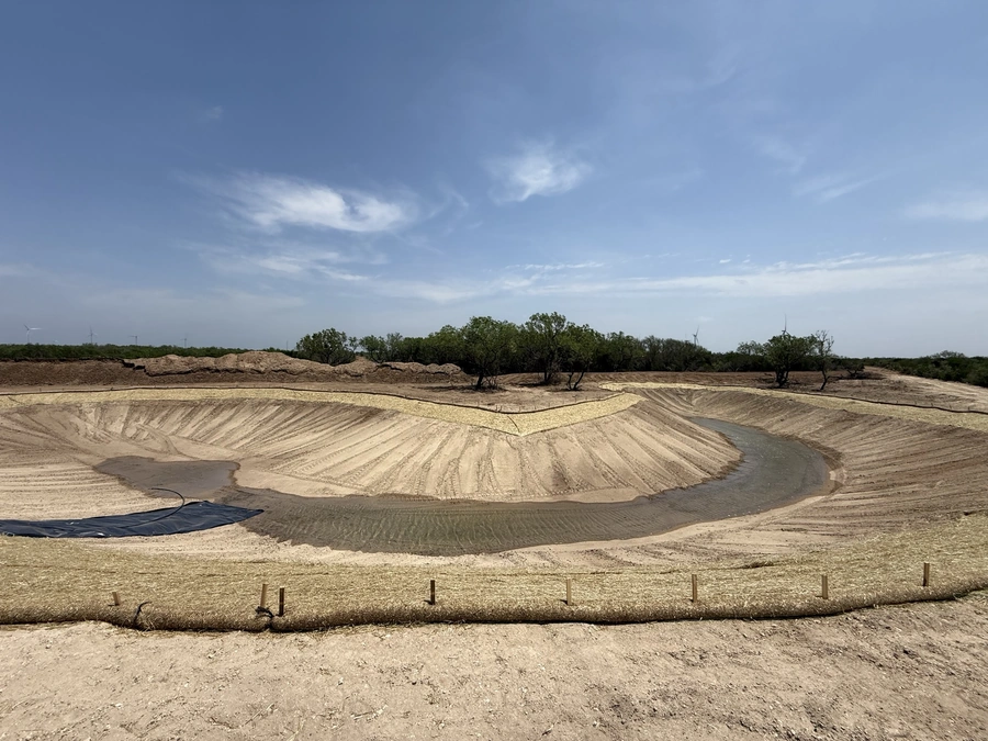 Aerial view of a large construction site with heavy equipment, earthworks, and a retention pond or pond being excavated. The site is surrounded by trees and vegetation, with a clear blue sky overhead.