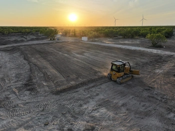 A yellow Caterpillar bulldozer working on a large, dusty construction site in a rural area. Rows of wind turbines are visible on the horizon against a vibrant sunset sky.