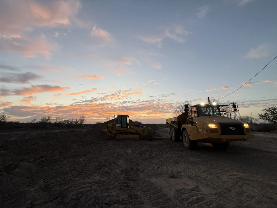 Two large construction vehicles, a bulldozer and a dump truck, working on a construction site at sunset. The sky is filled with vibrant orange and pink clouds, creating a dramatic backdrop for the scene.