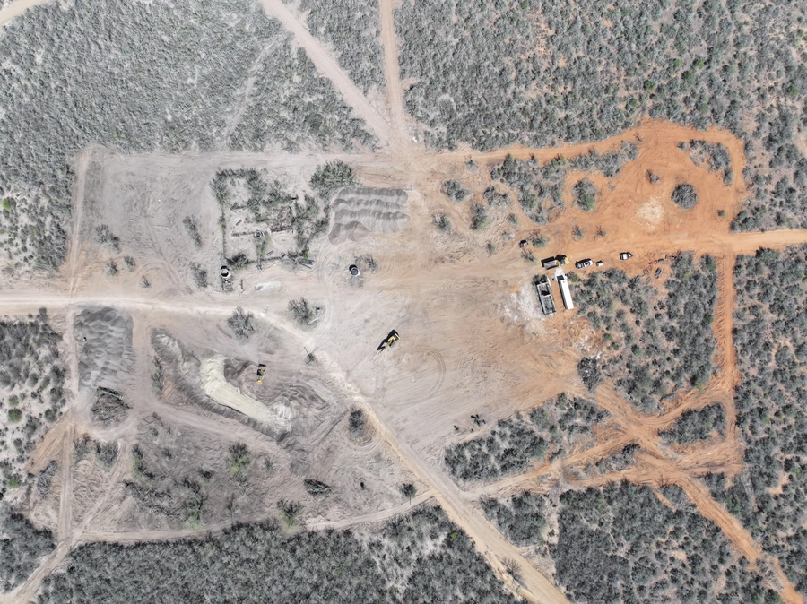 Aerial view of a construction site in a forested area, with various heavy machinery and buildings visible on the cleared land.