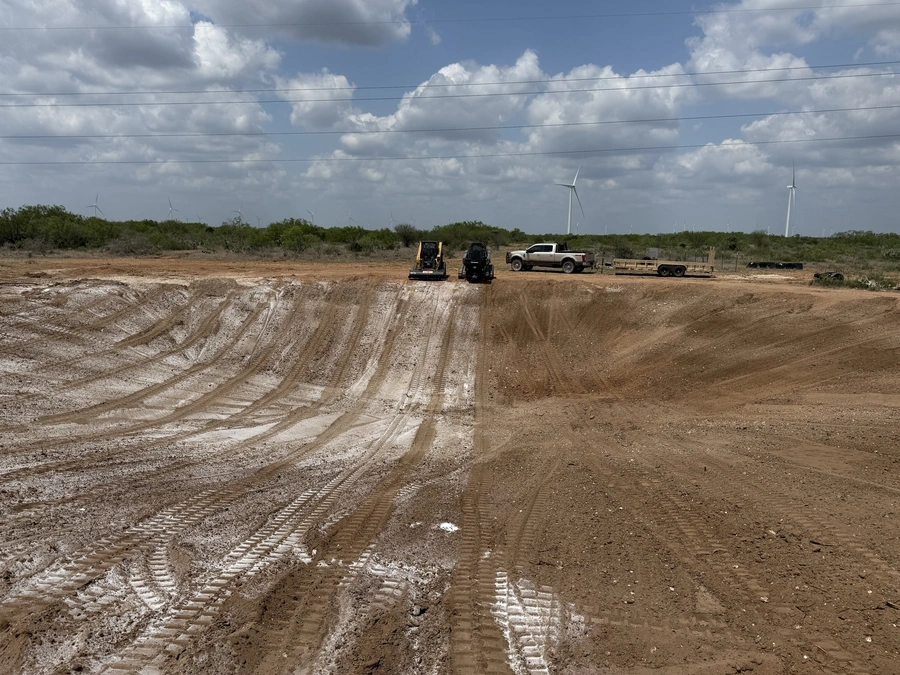 A construction site with heavy machinery, trucks, and wind turbines in the background. The scene shows excavation and land clearing work being done on a rural, open landscape.