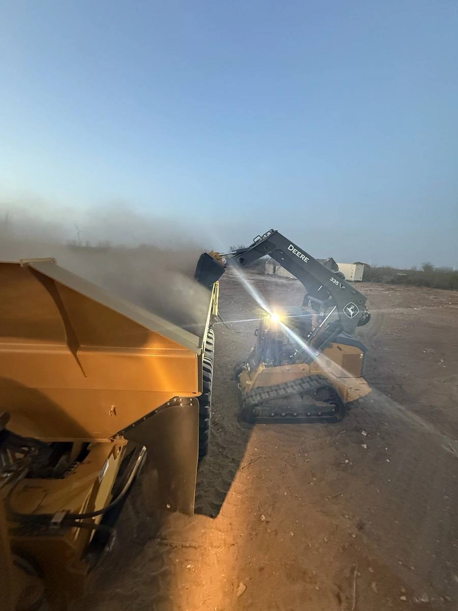 A Deere excavator machine clearing debris and dust on a construction site, kicking up a cloud of dust in the air.