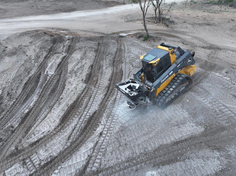 A yellow John Deere skid steer loader working on a dirt construction site, leaving tire tracks in the mud.