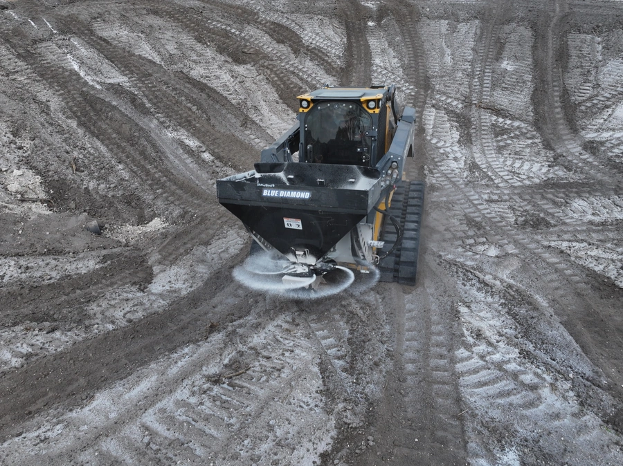 A blue tracked loader machine is clearing and grading a snowy construction site. The machine is leaving tracks in the snow as it moves across the uneven terrain.