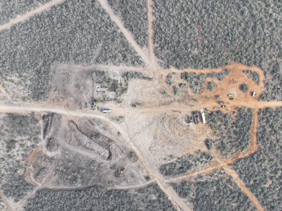 Aerial view of a construction site in a rural, forested area. The site shows extensive land clearing, earthworks, and partially constructed buildings or structures. Trucks and equipment are visible, indicating ongoing construction activities.