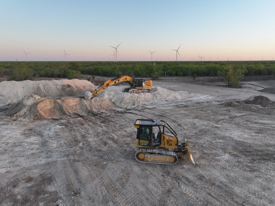 An aerial view of an active construction site with heavy machinery excavating and moving large piles of dirt. Wind turbines can be seen in the background, indicating this is a renewable energy project location.
