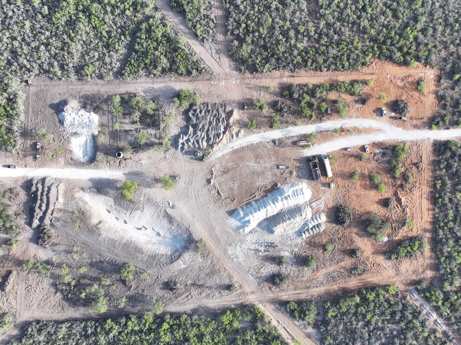 Aerial view of a construction site in a wooded area, with heavy machinery, excavated areas, and dirt piles visible.